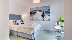 Un dormitorio moderno con una cama grande, una mesita de cristal con una botella y flores, y un cuadro de un faro en la pared.