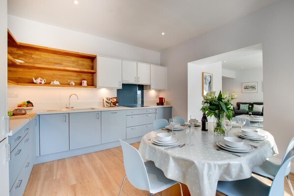 A modern kitchen with light grey cabinets, a wooden shelf, and a dining area with a round table set for four. - 3 Bed in St. Levan (oc-p00787) (Saint Levan)