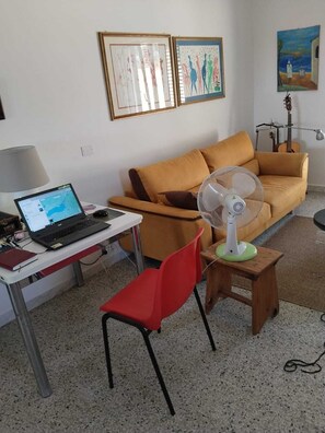 A room with a laptop on a desk, a red chair, a sofa, a standing fan, and two framed artworks on the wall. - Country house in an olive grove, 8 km from the Ionian Sea (Bernalda)