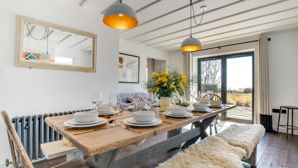 A dining area with a wooden table set for four, a fluffy bench, and a view of the outdoors. - 5 Bed in Walberswick (oc-lev) (Walberswick)