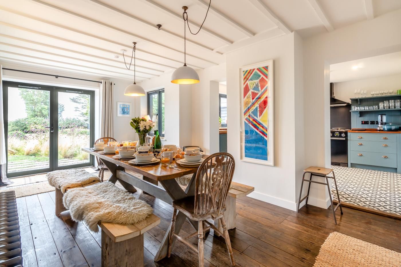 A dining area with a wooden table set for breakfast, a large window with a view of greenery, and a kitchen visible in the background.