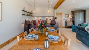 Un comedor con una mesa de madera dispuesta para seis personas, rodeada de sillas. En el fondo, se observa una cocina con electrodomésticos modernos y techo de madera.