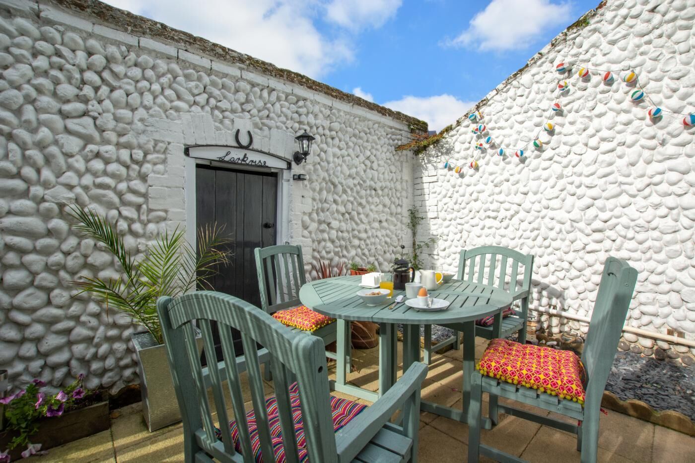 Un espacio al aire libre con paredes de piedra para comer, con una mesa, sillas, una puerta y una pared decorada con bolas de colores.