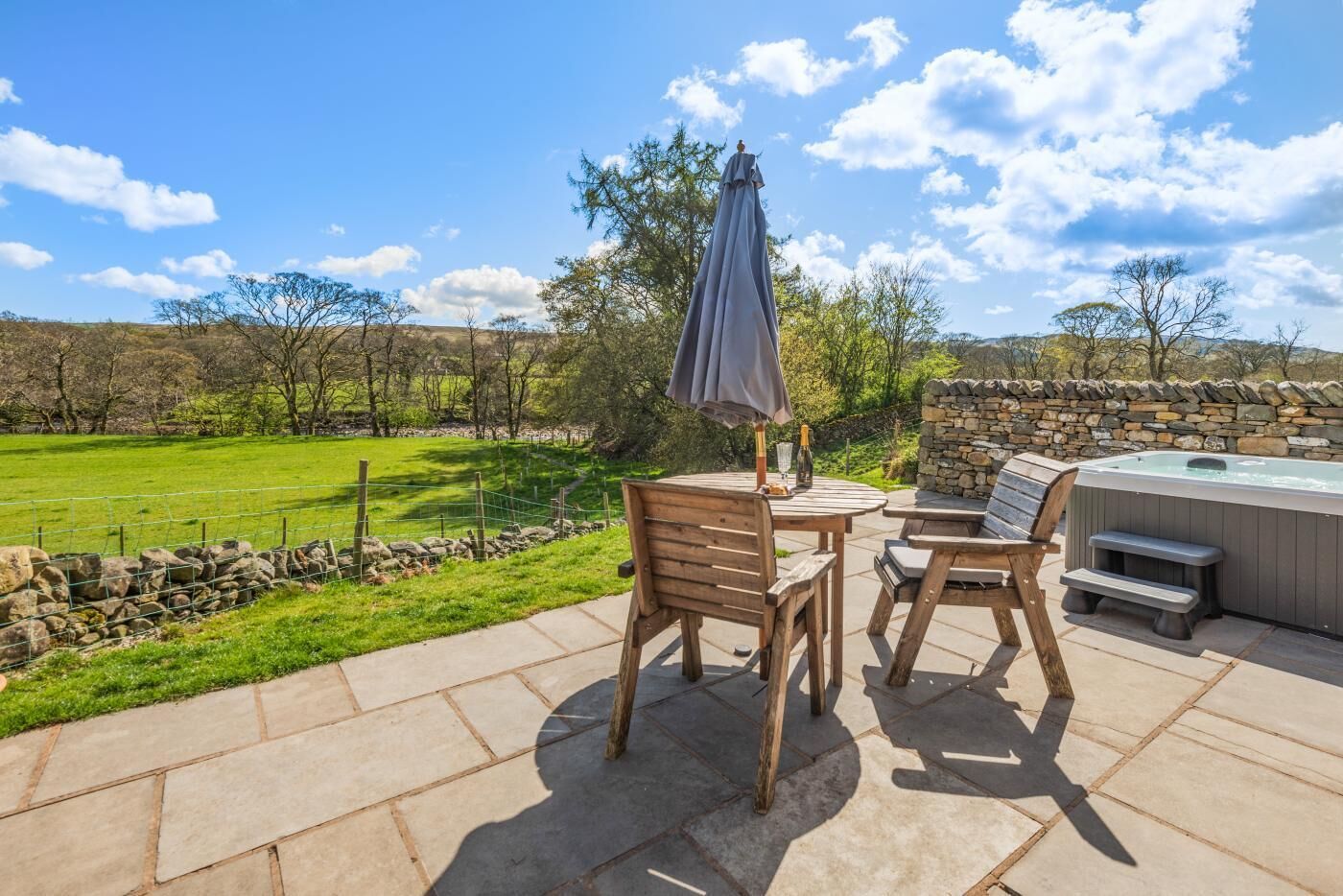 An outdoor patio with a stone table, two wooden chairs, a stone wall, and a hot tub.