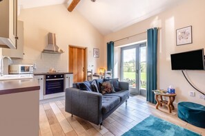 Eine moderne Küche mit Mikrowelle, Backofen und Holzschränken. Im Wohnbereich befindet sich ein graues Sofa mit Kissen, und ein Fernseher ist an der Wand montiert.