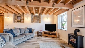 A cozy living room with a grey sofa, wooden beams, a TV, a fireplace, and stone walls.