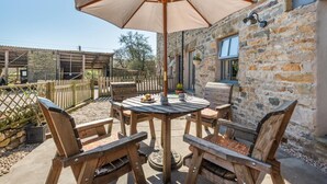 A rustic outdoor patio with a round wooden table and chairs, a large beige umbrella, and stone walls.