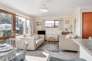 Un salon confortable avec un canapé, des fauteuils, une petite table et un téléviseur. Il y a des fenêtres avec des rideaux, une porte et une vue sur l’extérieur.