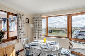 Un coin repas avec une table dressée pour quatre personnes, de grandes fenêtres avec des rideaux et une vue sur l’extérieur.