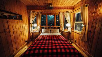 A wooden cabin bedroom with a checkered bedspread, two bedside lamps, and a window with curtains.