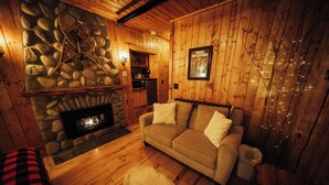 Un intérieur de chalet chaleureux avec une cheminée en pierre, des murs en bois, un canapé et un petit coin cuisine.