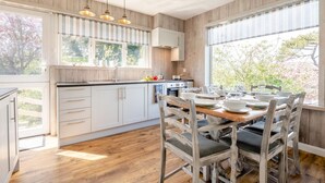 Una cocina moderna con armarios de madera, una zona de comedor con mesa y sillas de madera, y ventanas grandes con vistas a árboles.