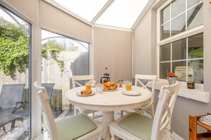A conservatory with a round table set for breakfast, including croissants and orange juice, surrounded by white chairs with green cushions. - 3 Bed in Hythe (oc-o31997) (Hythe)