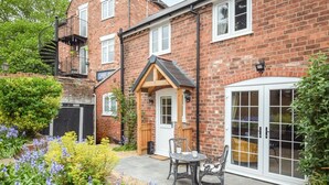 A brick house with a wooden front door, a small patio with a table and chairs, and a garden with various plants.