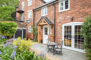 A brick house with a wooden front door, a small patio with a table and chairs, and a garden with various plants. - 2 Bed in Bewdley (oc-n31968) (Bewdley)