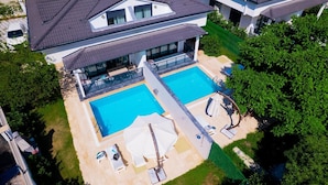 A modern two-story house with a large rectangular swimming pool, outdoor seating, and a well-maintained lawn.