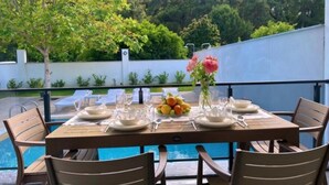 A dining table set outdoors with chairs, a vase with flowers, and a fruit bowl.