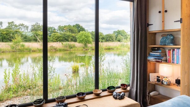 Ein Zimmer mit großem Fenster mit Blick auf einen Teich, einem Bücherregal mit Büchern und einem kleinen Tisch mit Schalen und einer Kamera.