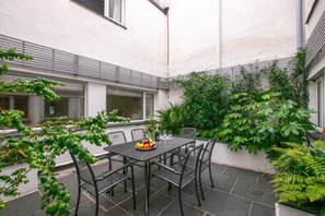 A patio with a table and chairs, surrounded by greenery and a white building. - 3 Bed in Padstow (oc-p00793) (Padstow)