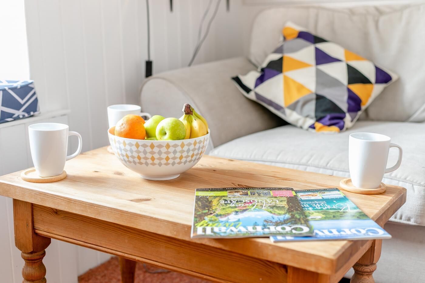 A wooden table with a bowl of fruit and two white cups, a magazine, and a patterned pillow on a couch.