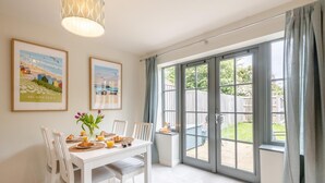 A dining area with a white table set for breakfast, featuring croissants and orange juice. There are two framed pictures on the wall, a large window with curtains, and a view of the backyard. - 2 Bed in Mundesley (oc-r31875) (Mundesley)