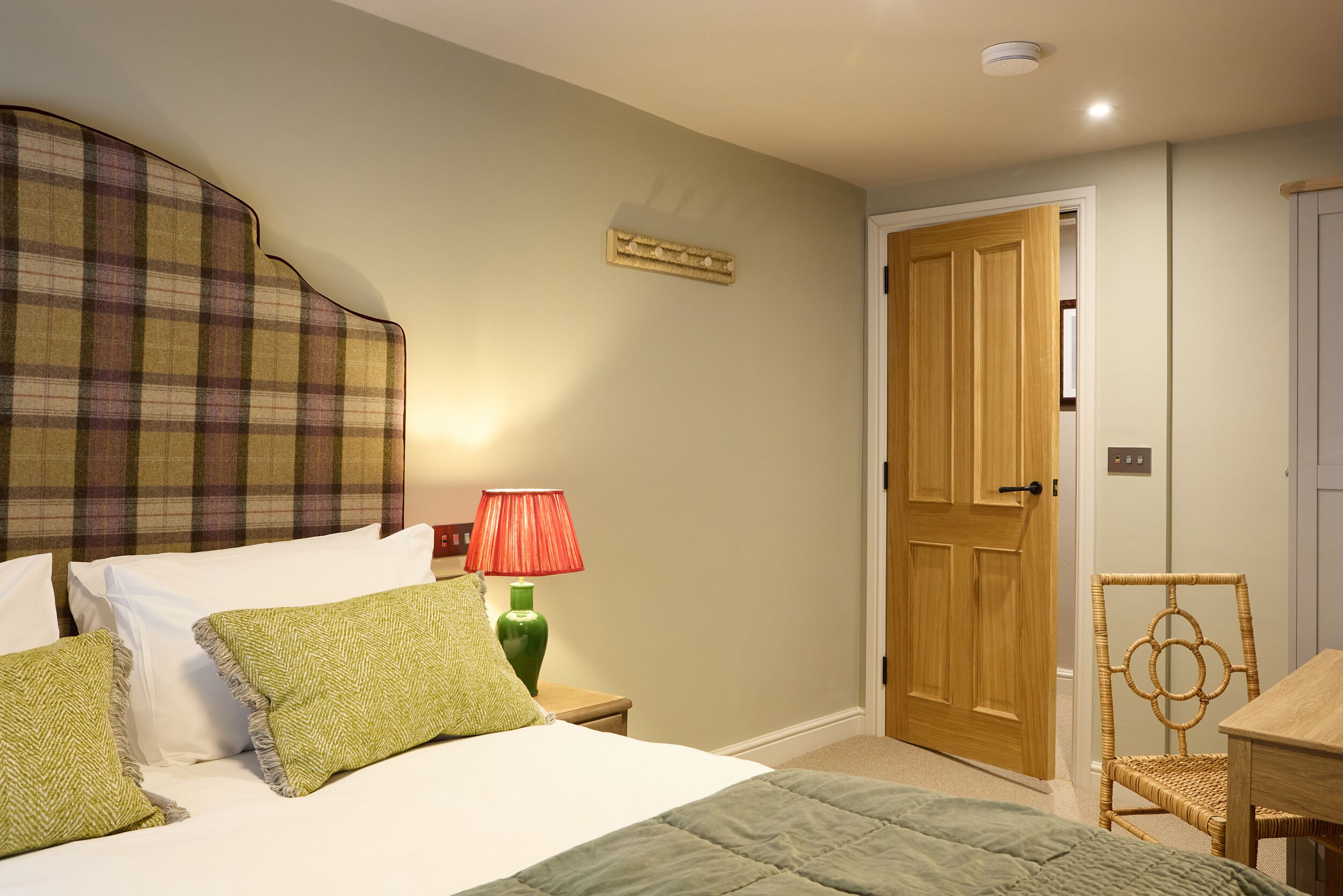 Une chambre à coucher avec un lit, des oreillers, une table de chevet, une lampe, une porte en bois et une chaise en osier.
