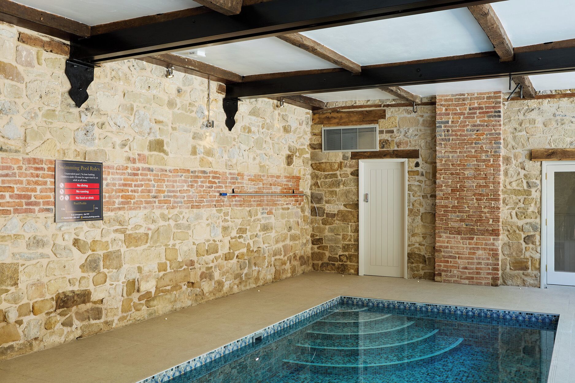 Una piscina con borde de azulejos, rodeada de muros de piedra y vigas de madera.