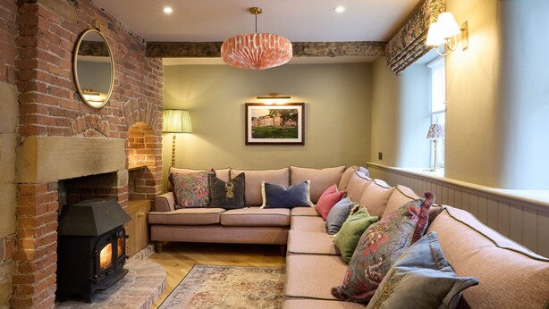 Una sala de estar acogedora con chimenea de ladrillo, un sofá con cojines coloridos, un cuadro enmarcado en la pared y una lámpara colgante.