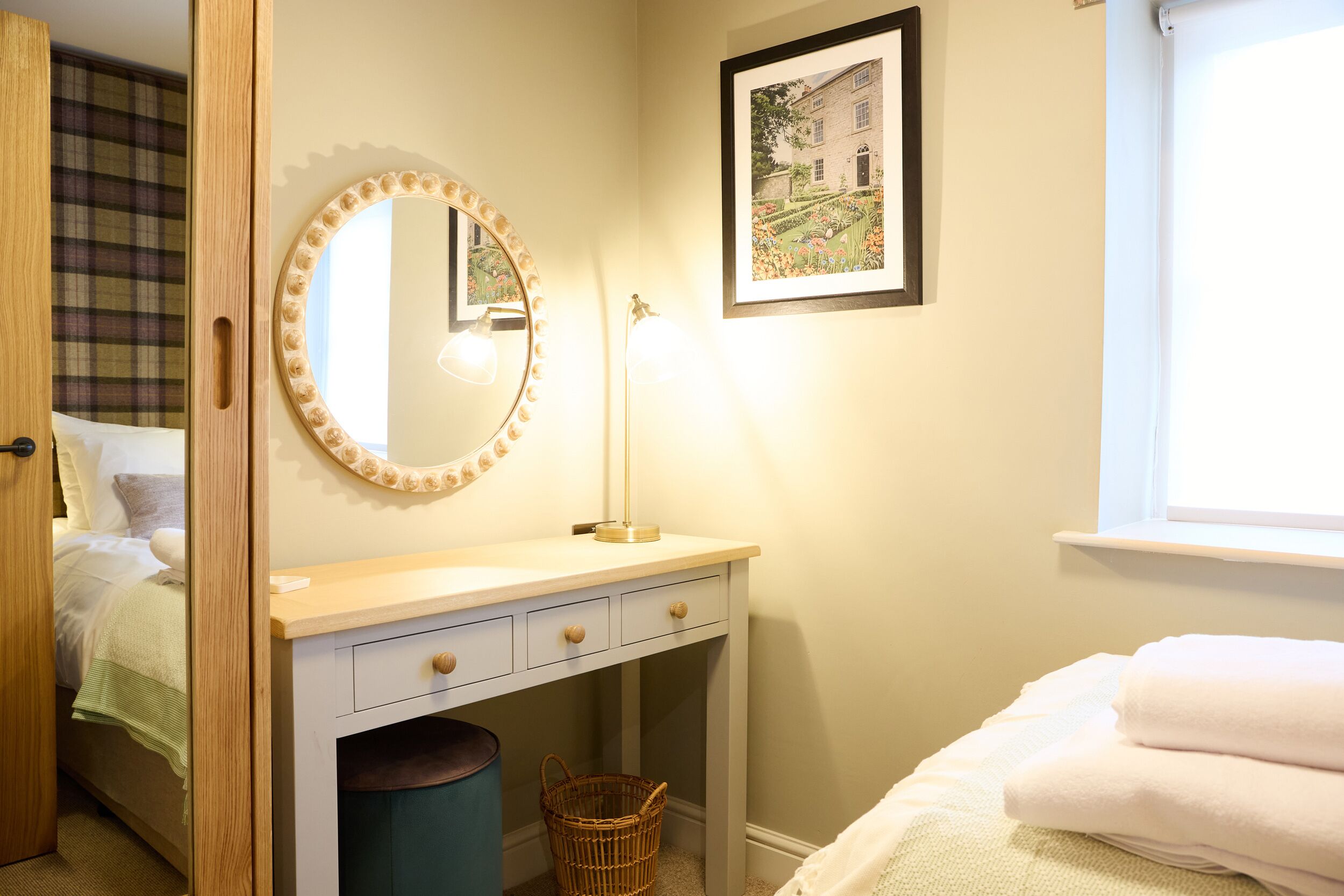 A bedroom with a bed, a bedside table with a lamp, a mirror, a framed picture on the wall, and a window.