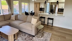 A modern living room with a sectional sofa, a small wooden table, and a glass coffee table. There is a kitchen area with a white countertop and bar stools. - Walk to Sabino Canyon Trails, 4Bd Home with Hot Tub (Tucson)
