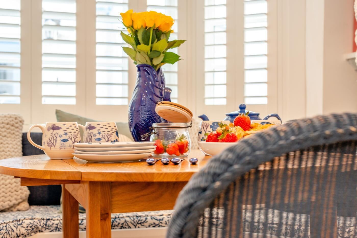A dining table with a vase of yellow roses, a glass jar with strawberries, and a bowl of fruit.