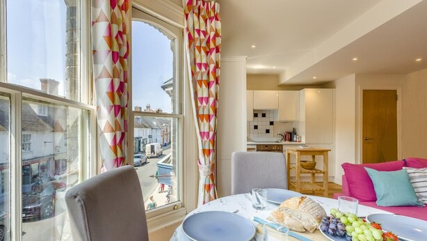 A dining area with a table set for two, a view of the street outside, and a kitchen in the background. - 1 Bed in Southwold (oc-bate) (Southwold)