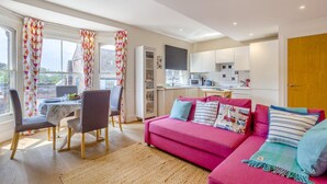 A modern living room with a pink sofa, a dining area with a table and chairs, and a kitchen with white cabinets. - 1 Bed in Southwold (oc-bate) (Southwold)