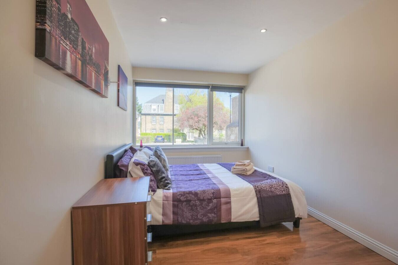 A bedroom with a large window, a bed with a quilt, a wooden dresser, and a painting on the wall.
