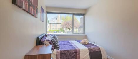 A bedroom with a large window, a bed with a quilt, a wooden dresser, and a painting on the wall.