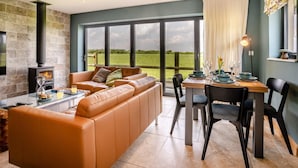 A modern living room with a stone fireplace, leather sofa, dining table, and large windows overlooking a green landscape.