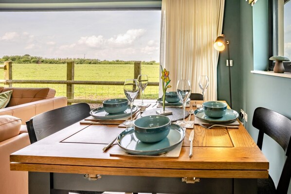 Una mesa de comedor servida con platos azules, vasos y cubiertos, con vista a un campo verde a través de una ventana.