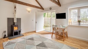 A room with a wood-burning stove, a dining table with chairs, a TV mounted on the wall, and a large window. - 1 Bed in Lamorna (oc-k31904) (Lamorna)