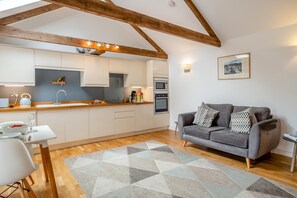 A modern kitchen with wooden beams, a dining area with a table and chairs, and a grey sofa with patterned cushions. - 1 Bed in Lamorna (oc-k31904) (Lamorna)