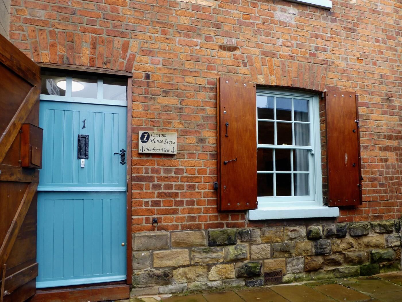 A brick building with a blue door and a window with wooden shutters. A sign on the wall reads "Custom House Steps, Harbour View."