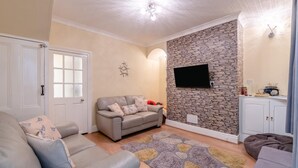 A living room with a grey sofa, a stone wall, a flat-screen TV, and a cabinet with a radio. - 2 Bed in Scarborough (oc-s027) (Scarborough)