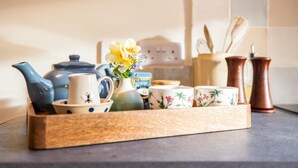 A wooden tray with a blue teapot, a white pitcher, floral cups, a vase with flowers, and two copper pepper shakers on a kitchen counter.