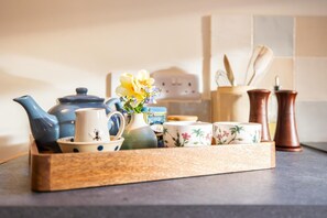 A wooden tray with a blue teapot, a white pitcher, floral cups, a vase with flowers, and two copper pepper shakers on a kitchen counter.