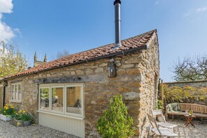 Una casa de piedra con techo de tejas, chimenea y un pequeño jardín con asientos.