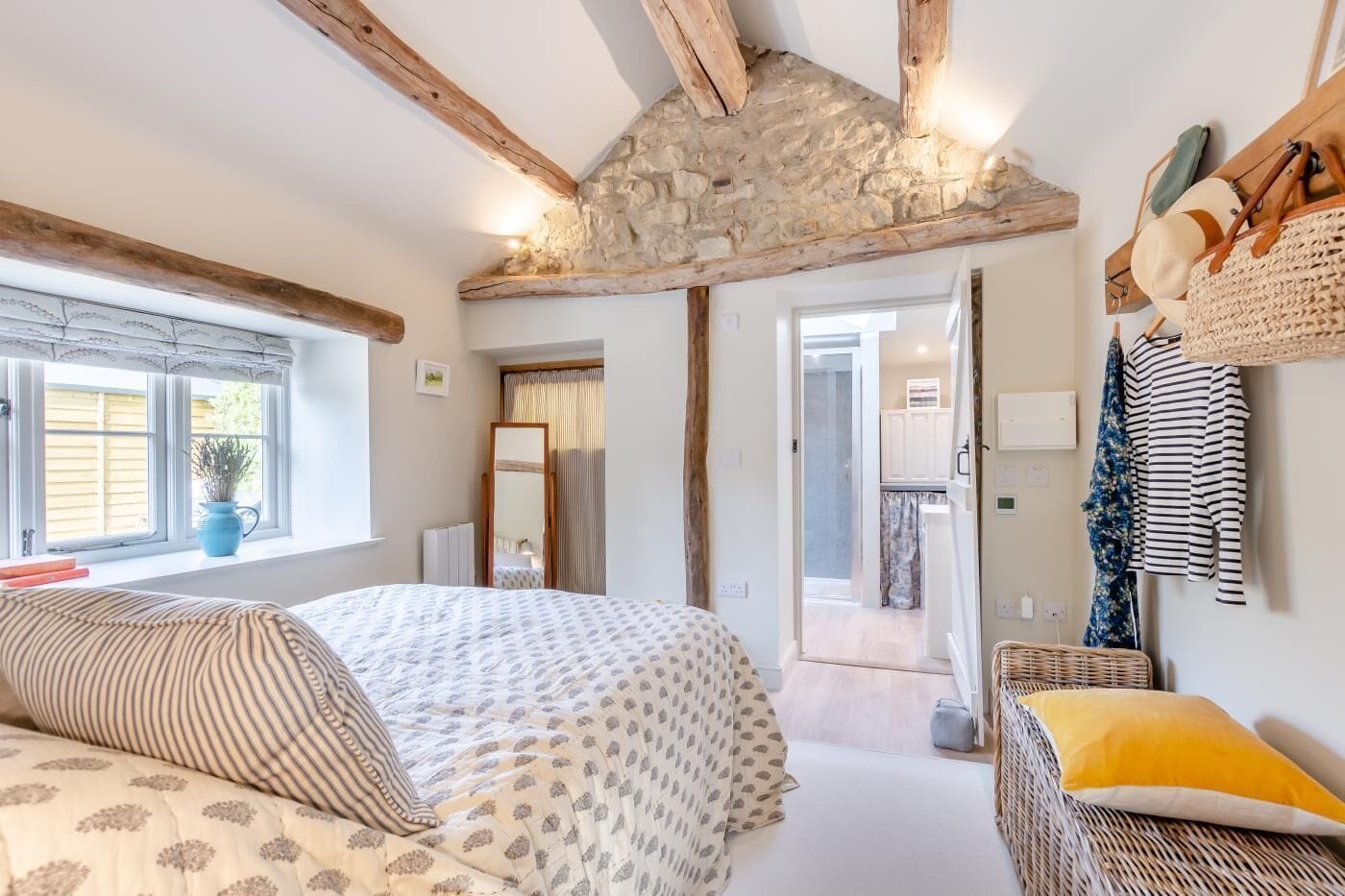 A bedroom with a large bed, a stone ceiling, wooden beams, a window with curtains, and a view into the kitchen.