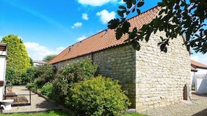 Un bâtiment en pierre surmonté d’un toit de tuiles, entouré d’un jardin bien entretenu et sous un ciel d’un bleu limpide.