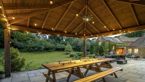 En overdækket terrasse med et træbord og bænke, omgivet af en have og en murstensbygning.