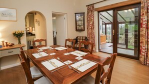 Un coin repas avec une table en bois dressée pour six personnes, entourée de chaises. Un lustre est suspendu au plafond, et une grande fenêtre avec des rideaux est visible.