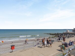 Eine Uferpromenade mit Kiesstrand, einem Rettungsschwimmturm und einer Mülltonne.