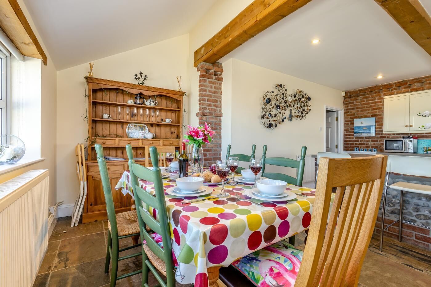 A dining area with a table set for four, surrounded by wooden chairs, a brick wall, and a kitchen area with a microwave.